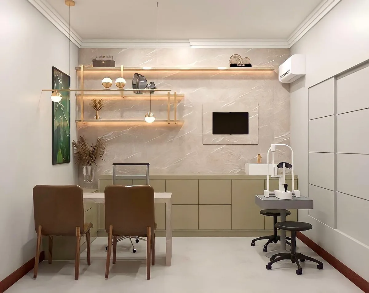 Consultório claro com mesa, cadeiras de couro, prateleiras com luz e equipamento oftalmológico.